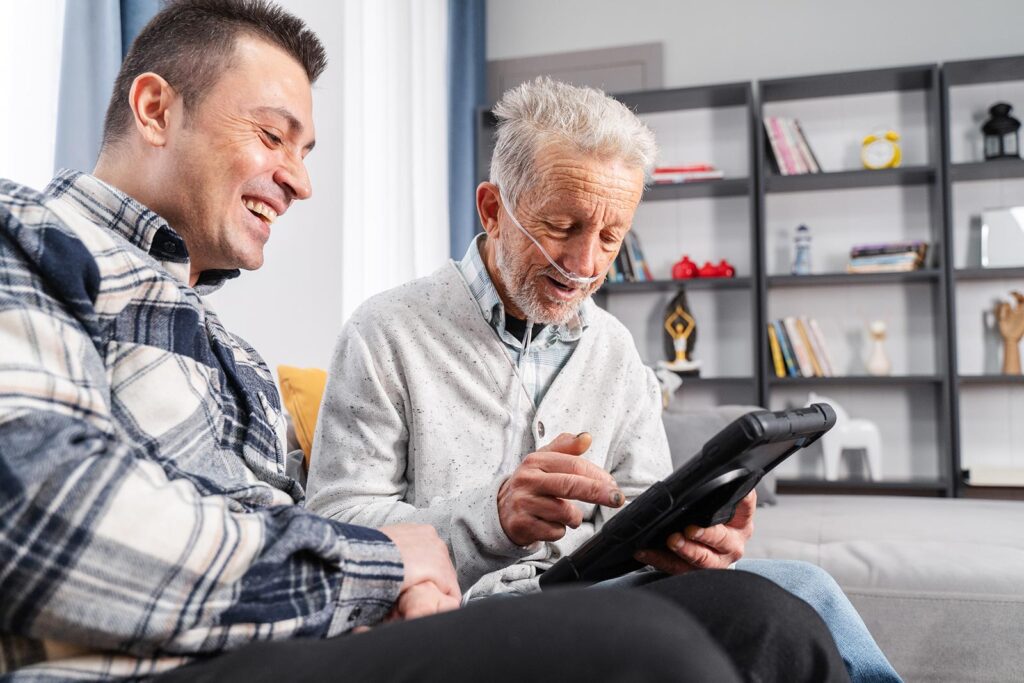 An older man on oxygen smiles with a family member as he manages his health on an tablet