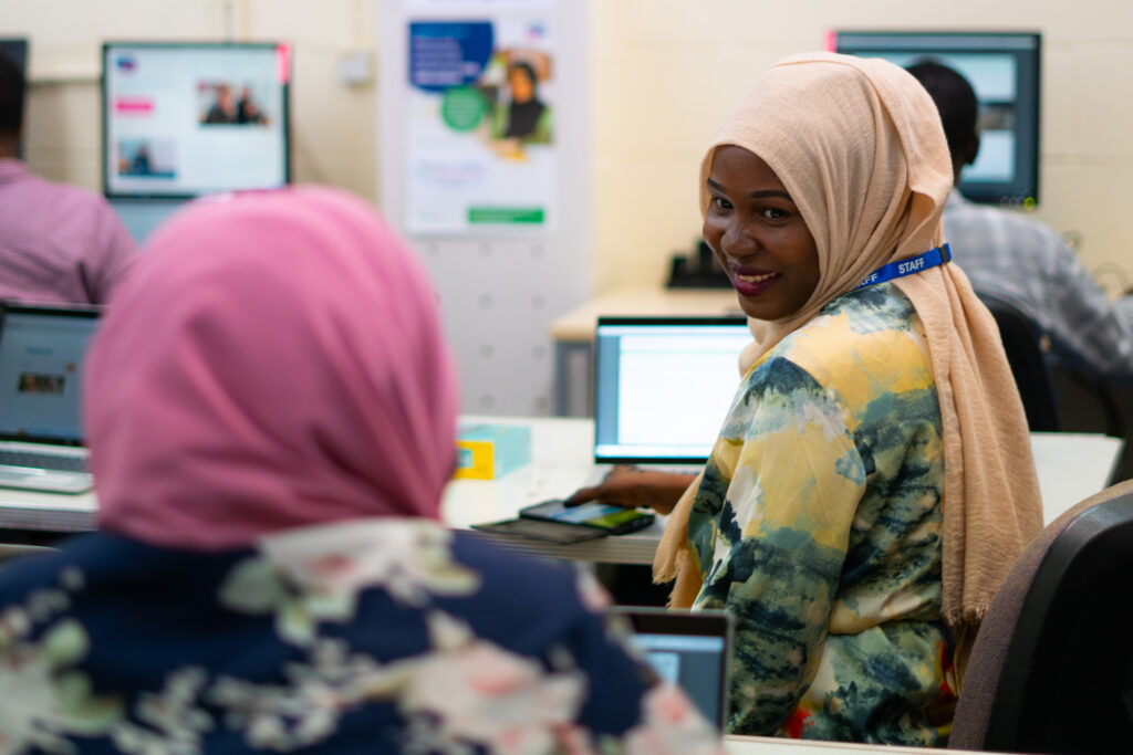 A migrant women training another in digital skills smiles at her student